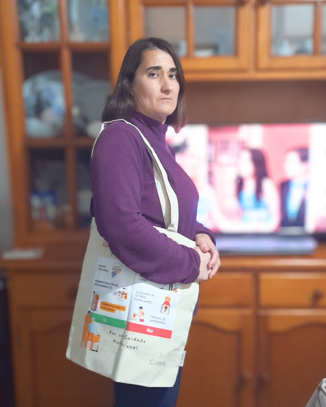 Mujer de pelo castaño, con jersey morado, posa con la totebag de Cuidarly en el salón de su casa.