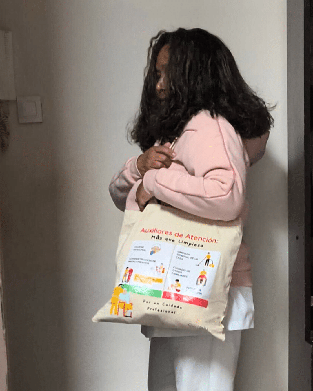 Mujer con sudadera rosa y cabello suelto muestra la totebag de Cuidarly en una habitación iluminada.