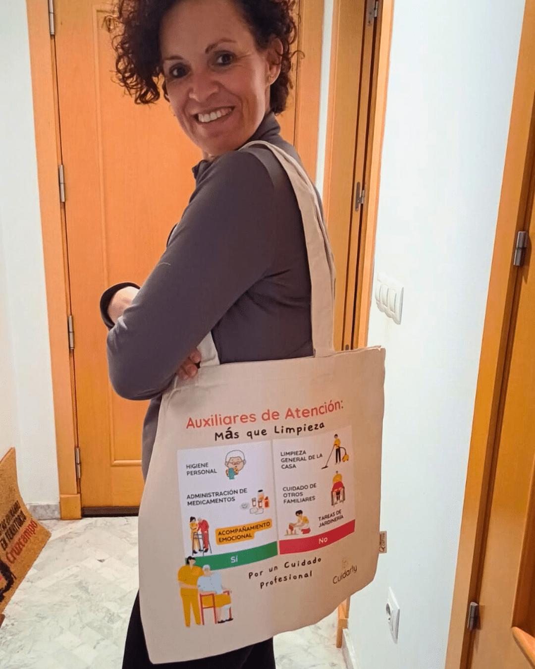 Mujer sonriente con pelo rizado lleva colgada al hombro la totebag de Cuidarly en el pasillo de su casa.