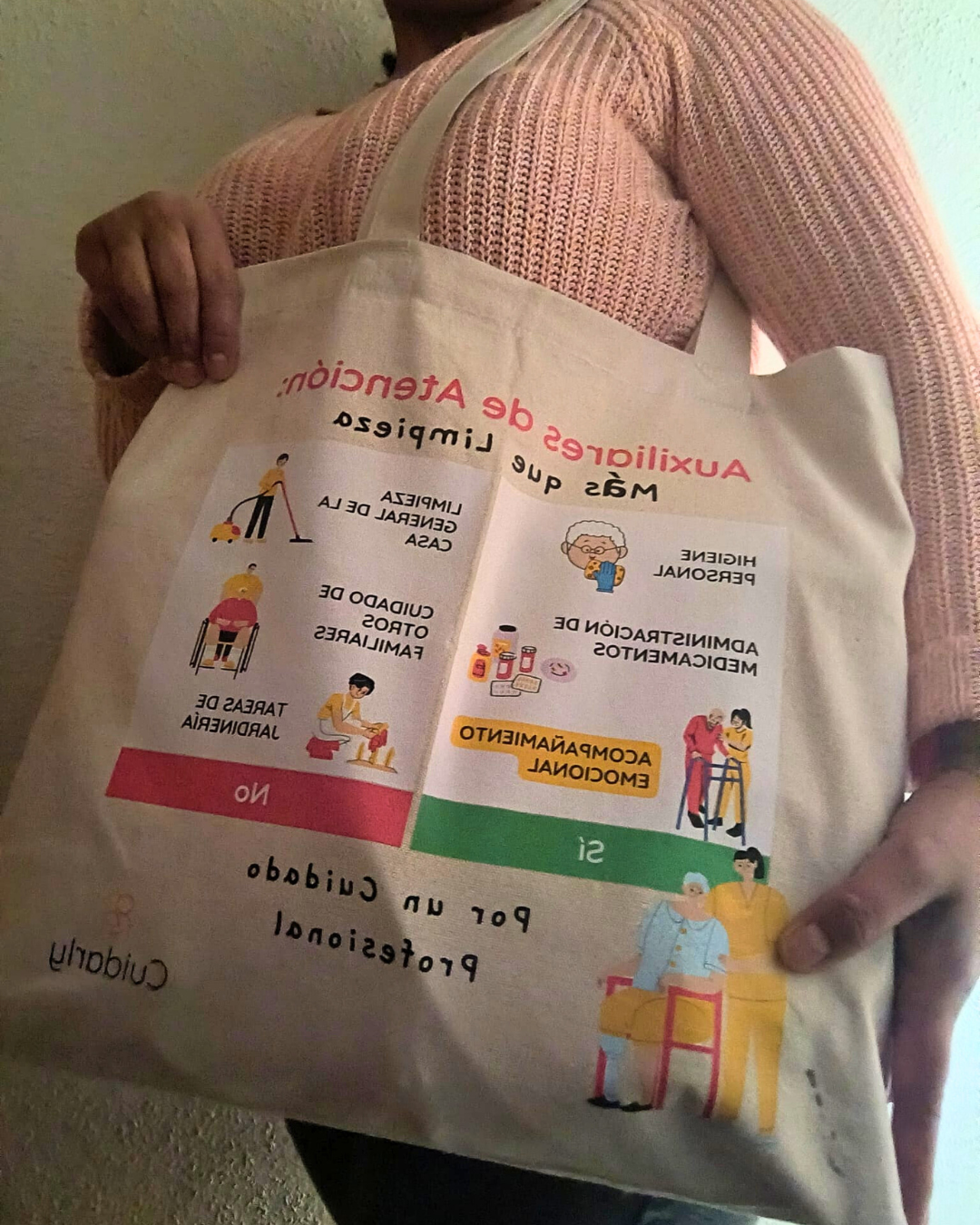Mujer con jersey rosa sosteniendo en primer plano la totebag de Cuidarly que reivindica que el trabajo de las auxiliares es más que limpiar.