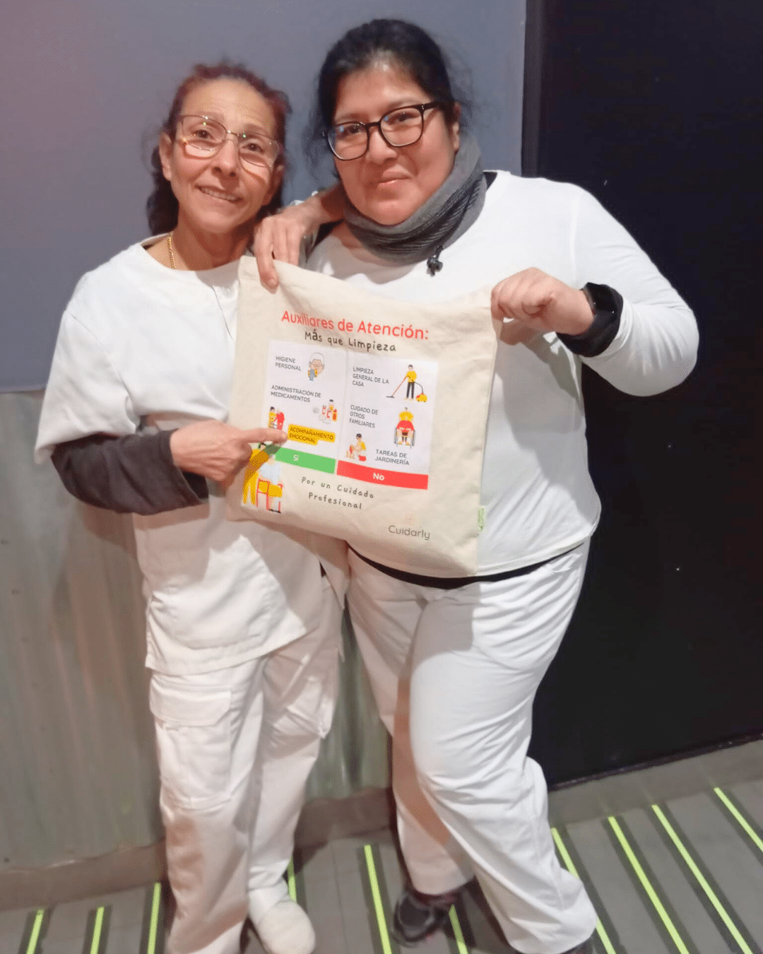 Dos mujeres auxiliares de ayuda a domicilio con uniforme blanco, una de ellas señalando la totebag de Cuidarly con el mensaje "Auxiliares de atención: más que limpieza".