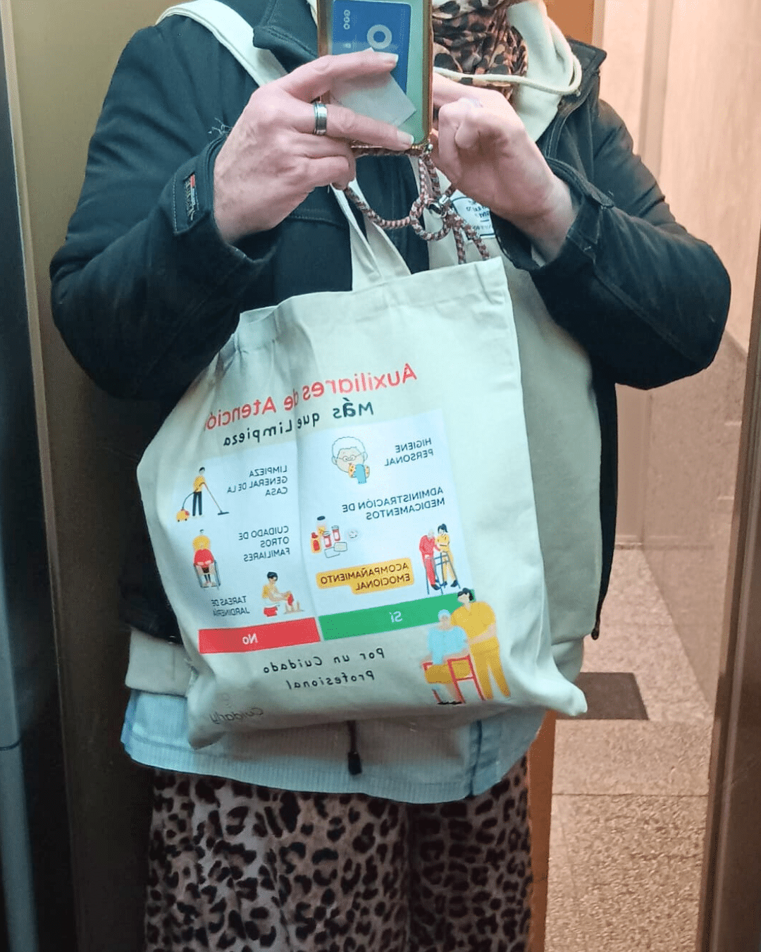 Auxiliar fotografiada dentro de un ascensor, mostrando su totebag de Cuidarly con el mensaje "Más que limpieza", parcialmente visible entre su abrigo y mochila.