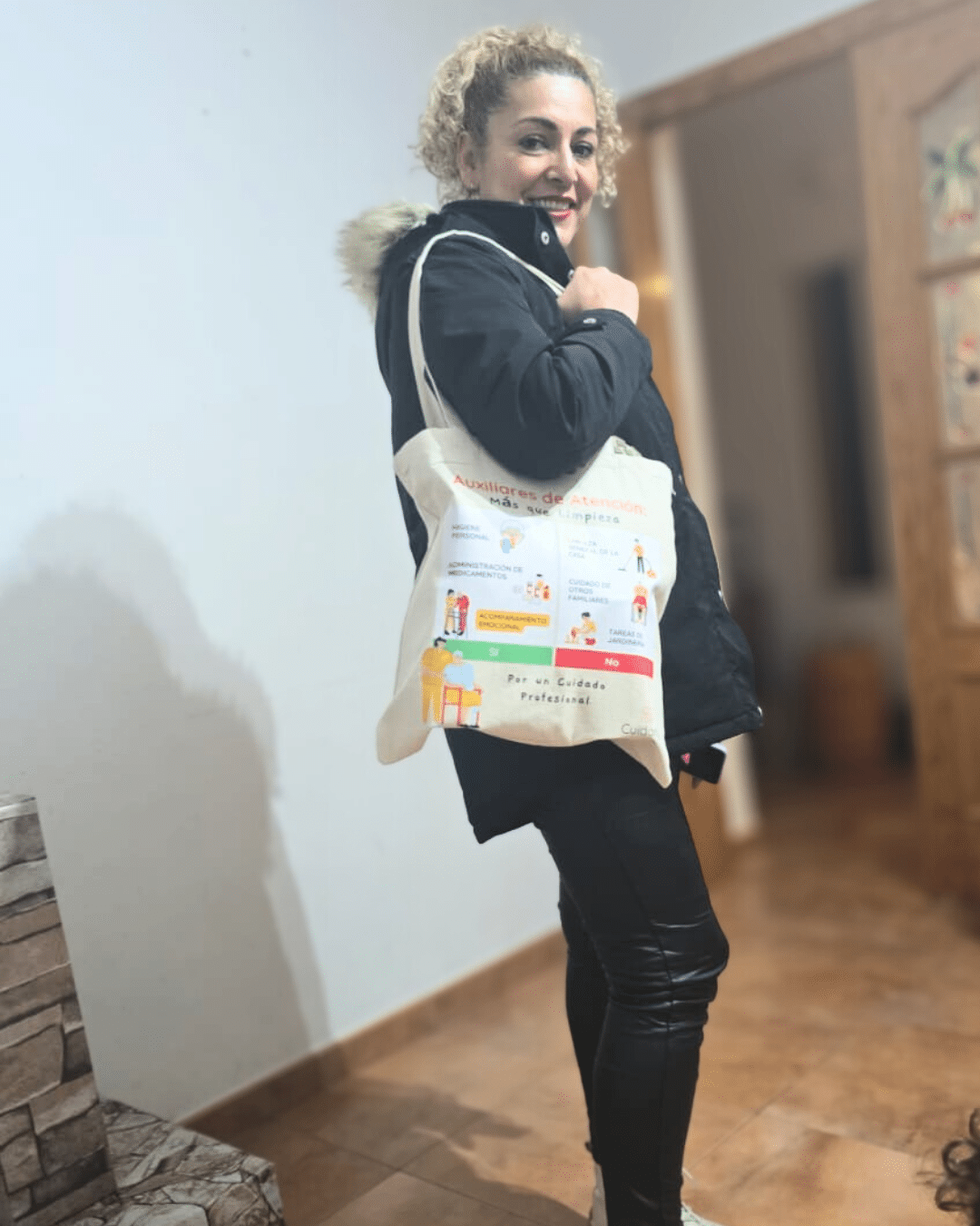 Auxiliar de ayuda a domicilio con abrigo negro y pantalón de cuero mostrando orgullosa su totebag Cuidarly con el diseño informativo sobre funciones profesionales.