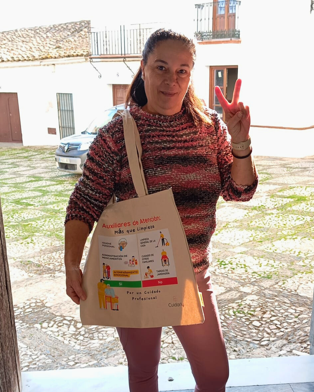 Auxiliar de atención domiciliaria sonriendo y haciendo el gesto de la victoria con los dedos mientras muestra la totebag de Cuidarly con el mensaje "Más que limpieza".