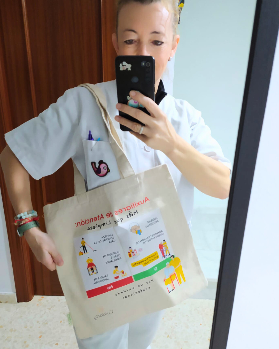 Auxiliar de ayuda a domicilio con su uniforme blanco, mostrando la totebag Cuidarly frente al espejo.