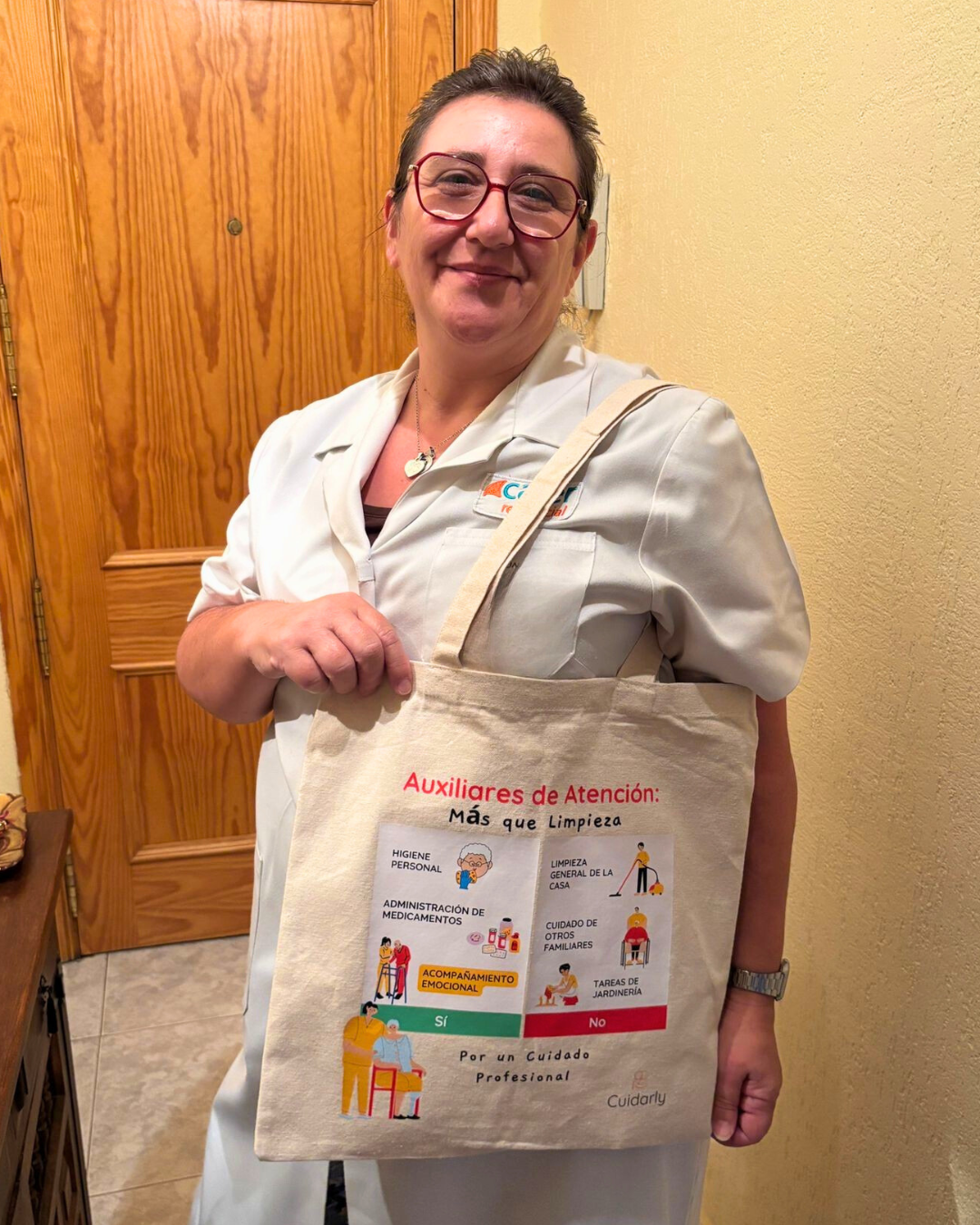 Auxiliar de ayuda a domicilio sonriendo con gafas y uniforme blanco, sosteniendo su totebag Cuidarly frente a una puerta de madera.