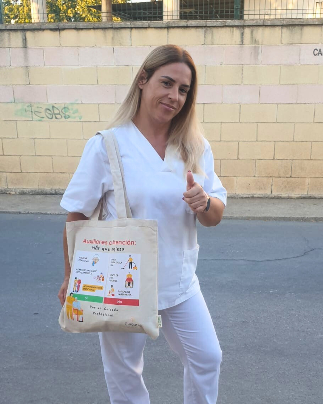 Auxiliar de ayuda a domicilio vestida de blanco, en la calle, mostrando la totebag de Cuidarly con el pulgar en alto.