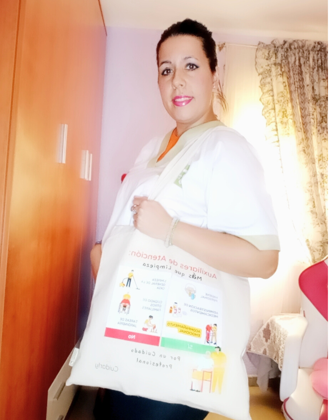 Auxiliar de ayuda a domicilio con uniforme blanco mostrando la totebag de Cuidarly en una habitación con cortinas florales.
