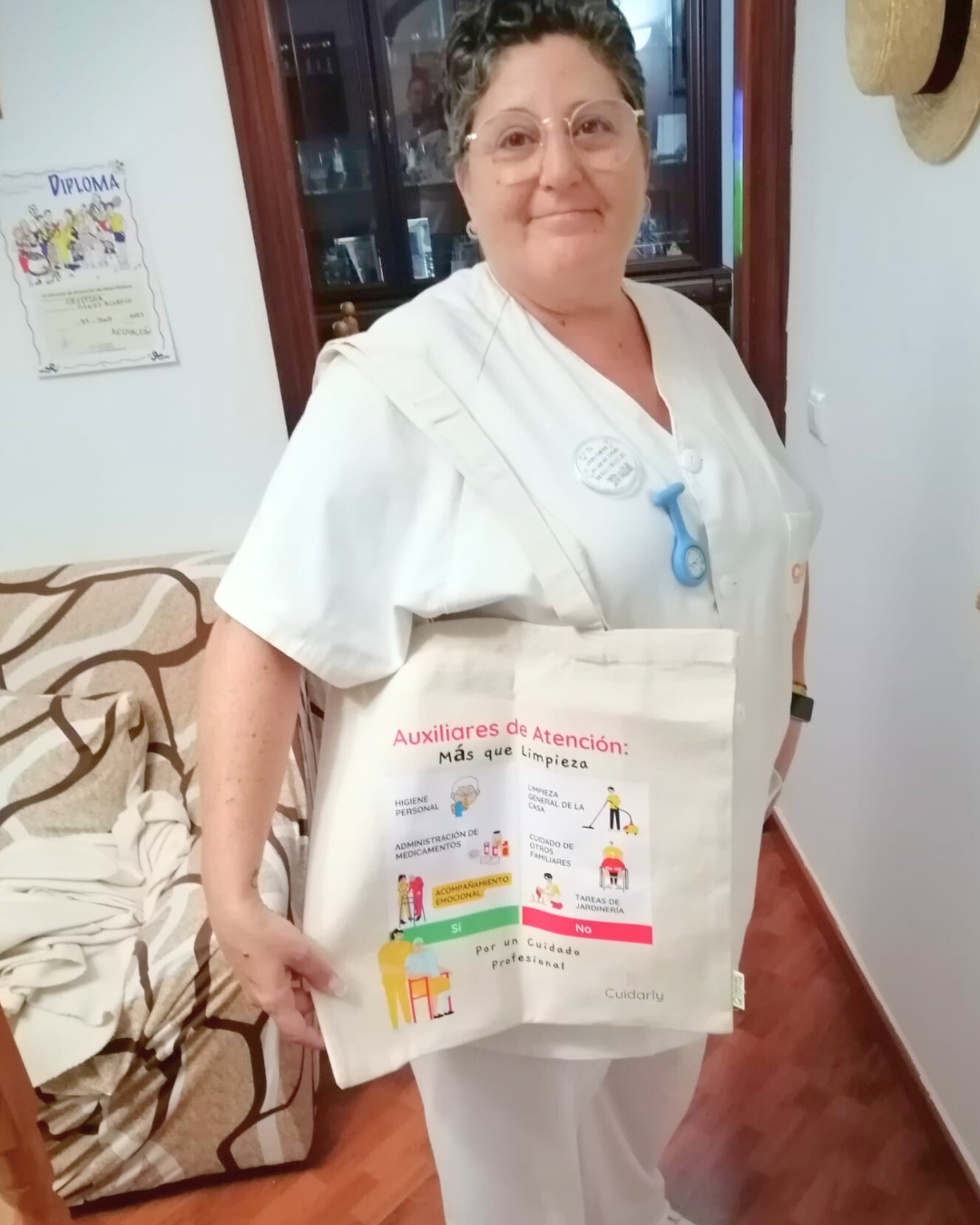 Mujer con uniforme blanco, gafas y totebag de Cuidarly posando con una expresión serena y orgullosa.